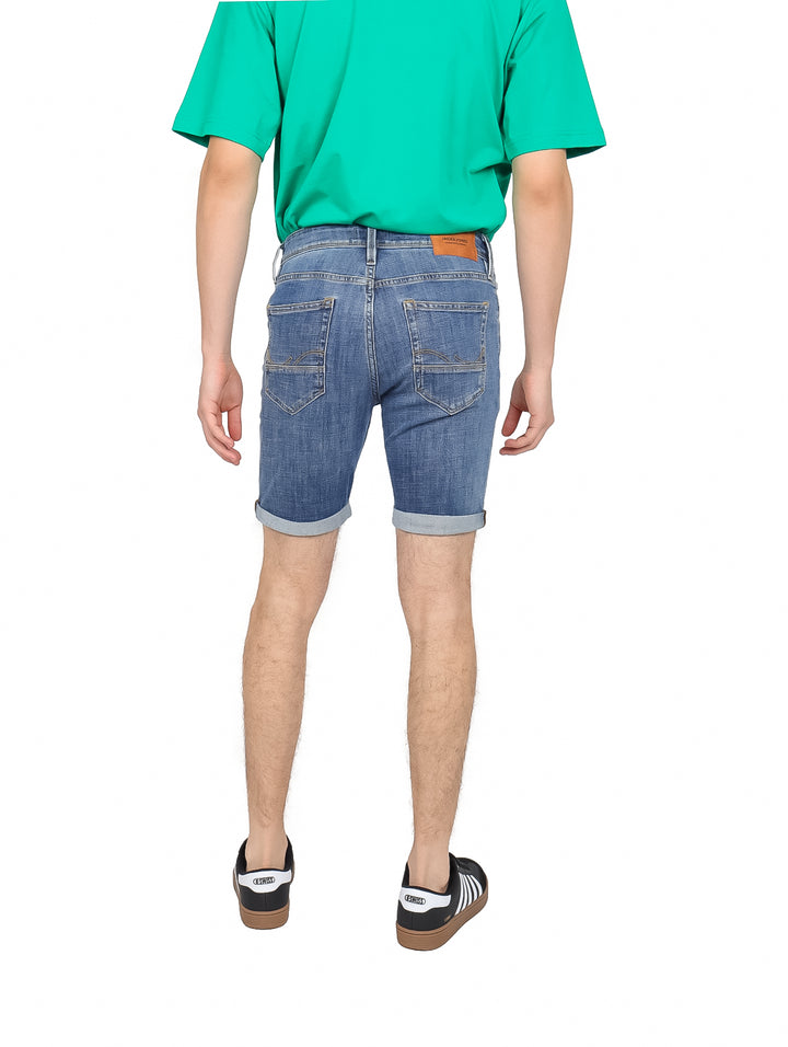 Hombre vistiendo shorts de mezclilla color azul, camiseta verde y tenis negros sobre fondo blanco.
