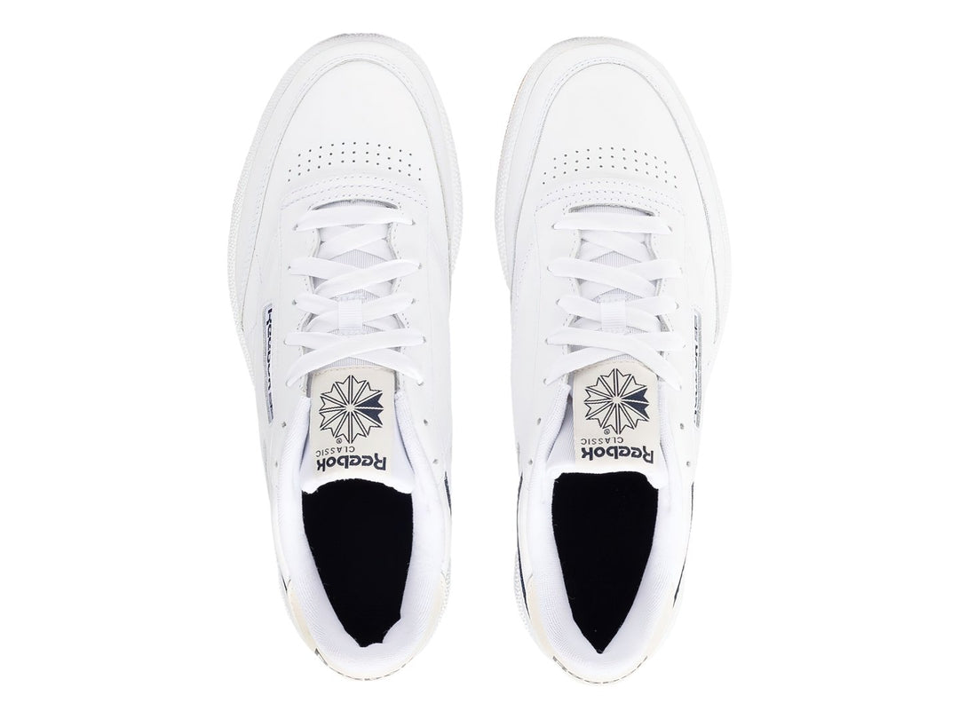 Tenis Reebok Club C 85 blancos con suela de goma color miel y detalle lateral en azul marino.