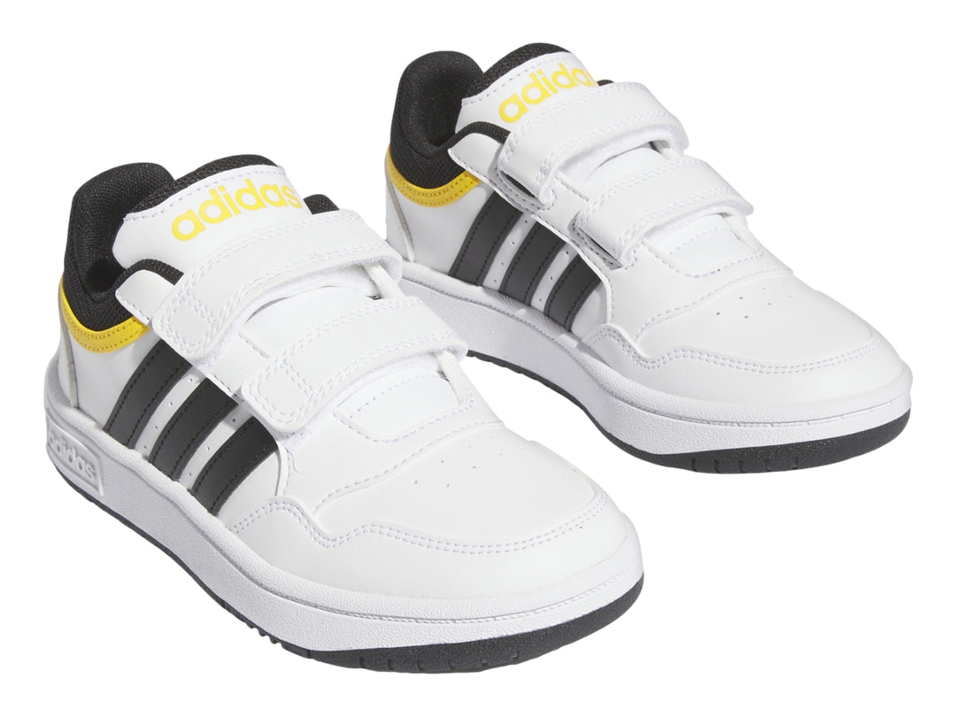 Tenis Adidas blancos con tres franjas negras, detalles amarillos y cierre de velcro.