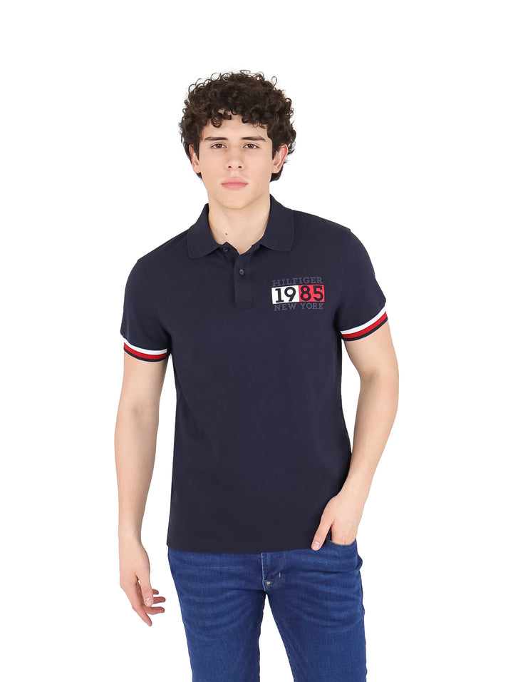 Hombre vistiendo polo Tommy Hilfiger color azul marino con detalles rojos y blancos en las mangas y logotipo en el pecho.