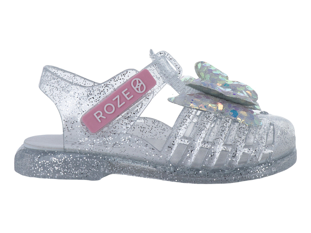 Sandalia infantil color plata con glitter y un moño iridiscente en la parte frontal.