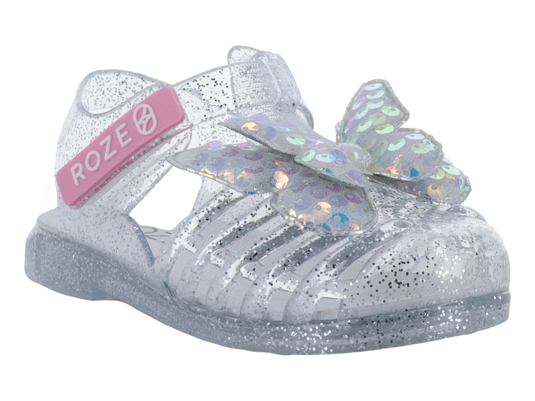 Sandalia infantil color plata con glitter y un moño iridiscente en la parte frontal.