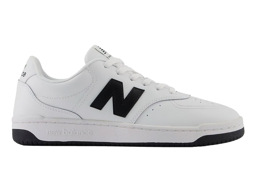 Tenis New Balance blancos con logotipo N en negro y suela negra, vista lateral.