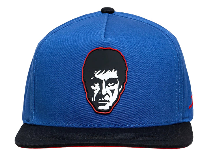 Gorra snapback azul con visera negra, presenta un bordado de rostro en el frente y logo lateral rojo.