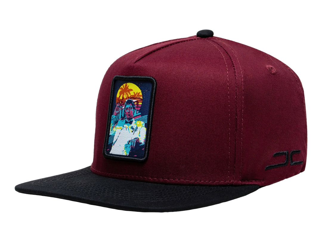 Gorra snapback color burgundy con visera negra y un parche rectangular colorido en el frente.