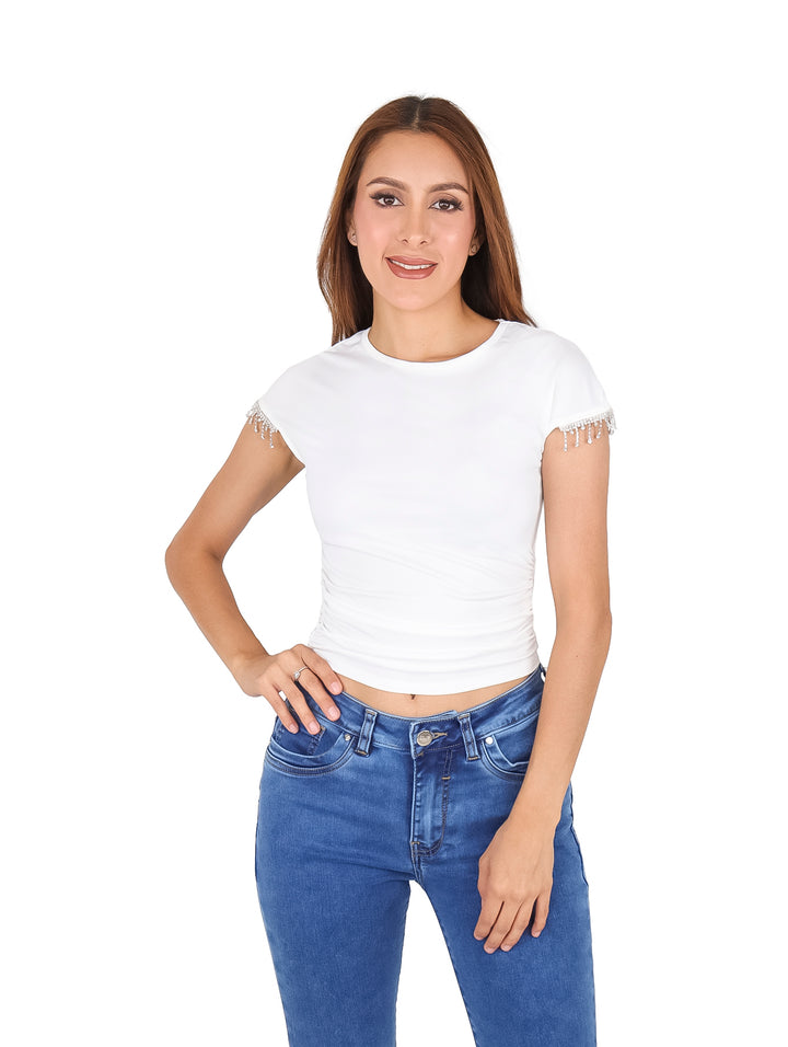 Mujer vistiendo blusa blanca de manga corta con detalles de piedras brillantes en las mangas y jeans azules.