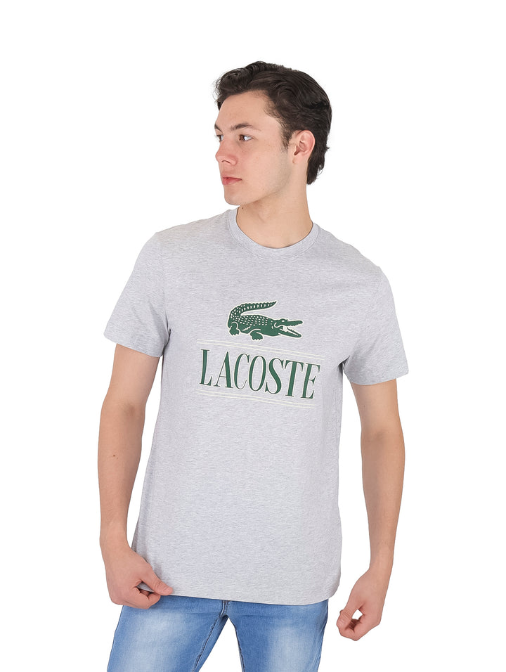 Hombre vistiendo una camiseta de manga corta color gris claro con logotipo de Lacoste en el pecho.