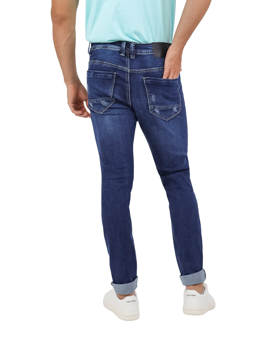 Hombre vistiendo pantalones jeans de mezclilla azul oscuro con detalles desgastados y tenis blancos.