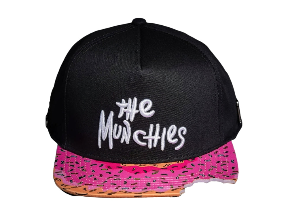 Gorra snapback negra con visera rosa estampada y bordado blanco que dice 'The Munchies'.
