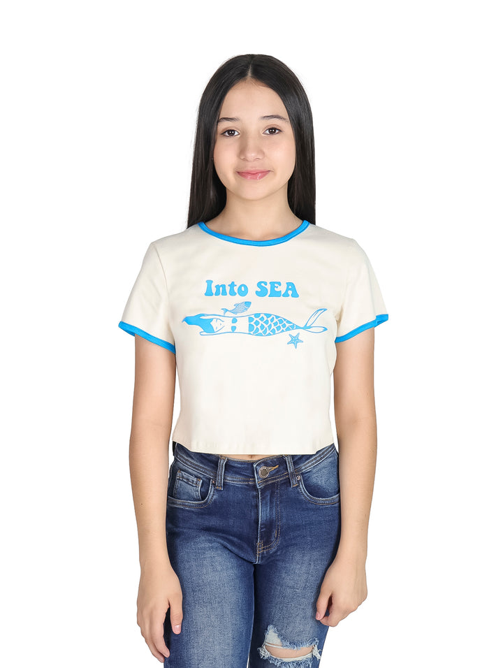 Niña vistiendo camiseta corta color crema con estampado de sirena y bordes en color azul.