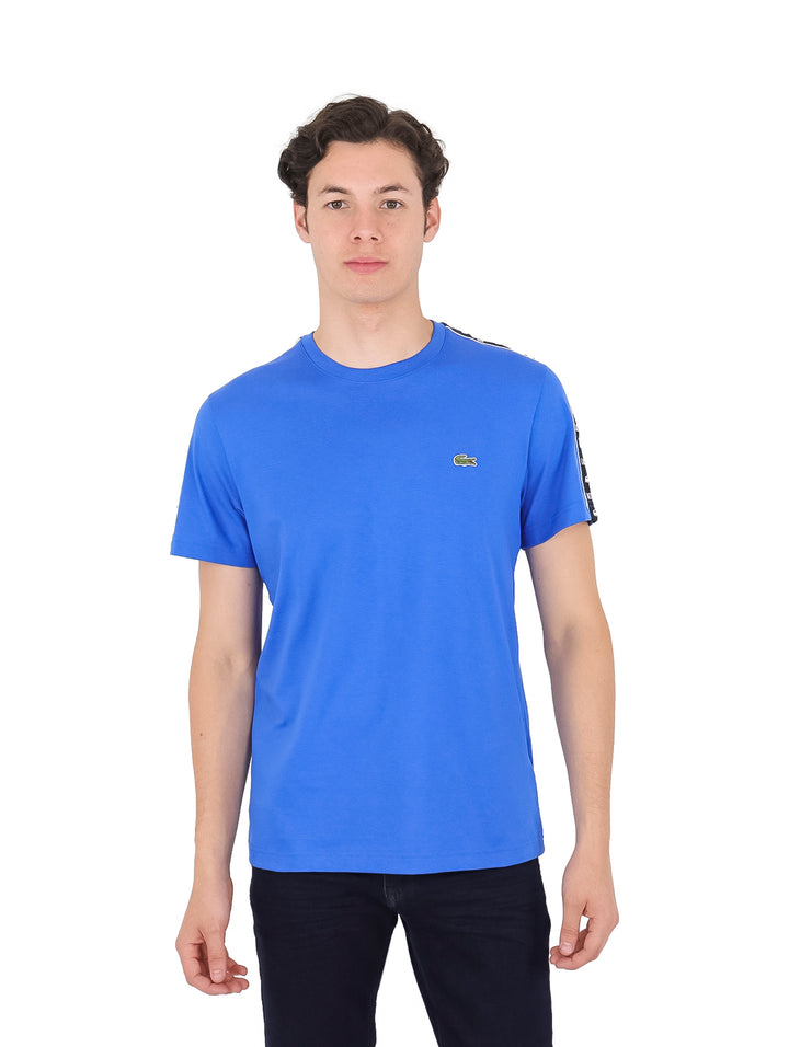 Hombre vistiendo camiseta azul de manga corta marca Lacoste con cintas decorativas negras en los costados.