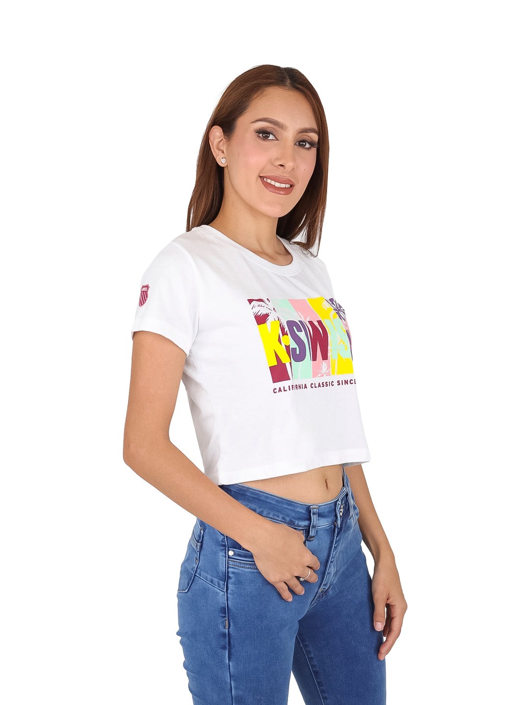Mujer vistiendo playera blanca crop top con logotipo colorido de K-Swiss y pantalones azules.