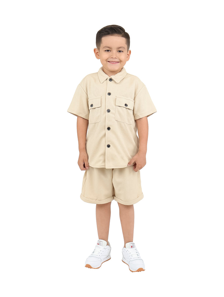 Niño vistiendo un conjunto casual de camisa y short en color beige con tenis blancos.