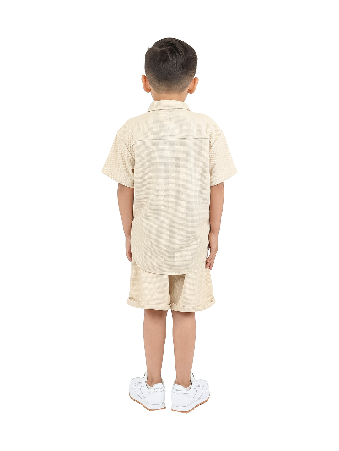 Niño vistiendo un conjunto casual de camisa y short en color beige con tenis blancos.