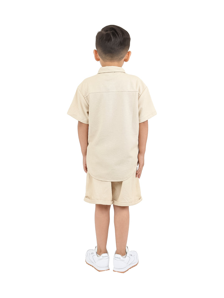 Niño vistiendo un conjunto casual de camisa y short en color beige con tenis blancos.