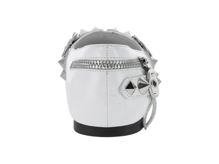 Zapato plano blanco con punta afilada, adornado con cierres y estoperoles plateados.