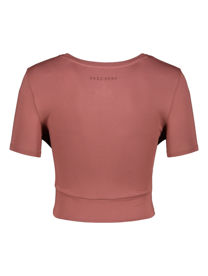 Mujer vistiendo playera deportiva Skechers color rosa oscuro con diseño cruzado en la cintura.