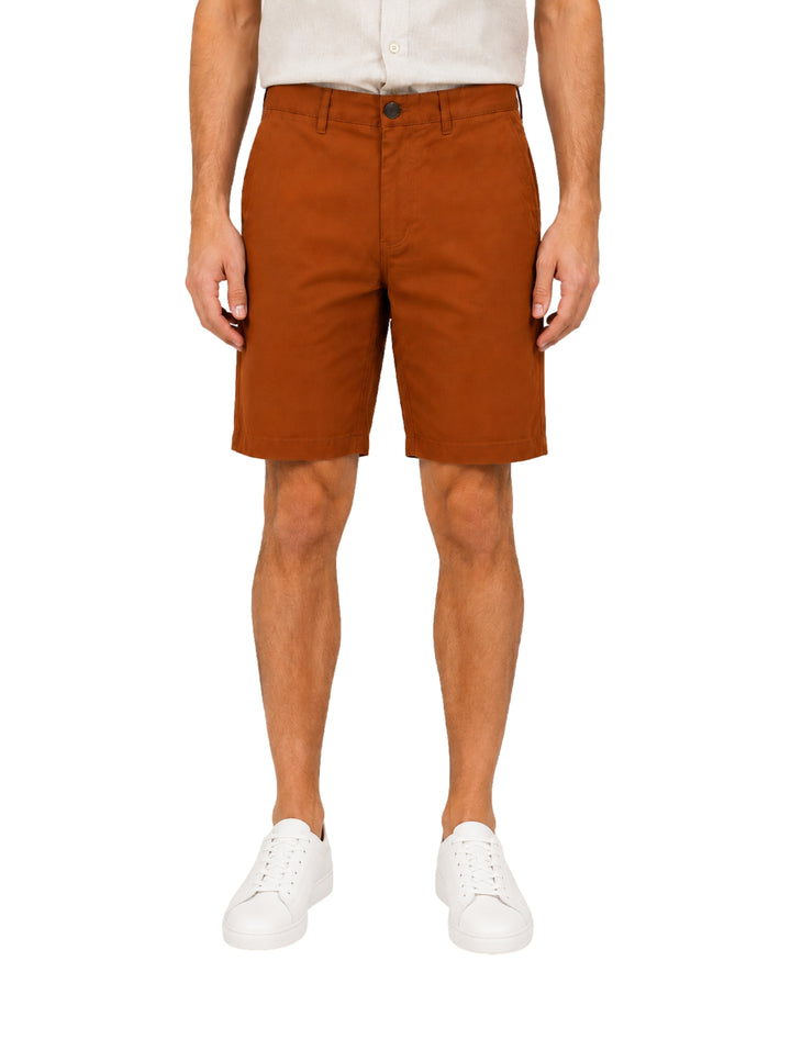 Hombre vistiendo shorts color tabaco con camisa blanca y tenis blancos sobre fondo blanco.