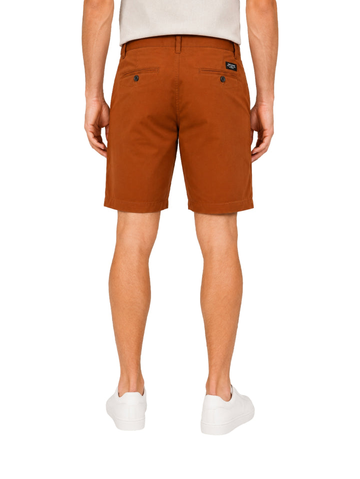 Hombre vistiendo shorts color tabaco con camisa blanca y tenis blancos sobre fondo blanco.