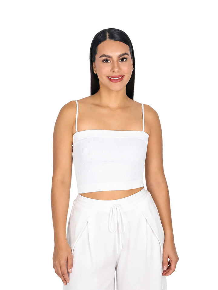 Mujer vistiendo blusa blanca corta de tirantes y pantalones blancos sobre fondo blanco.