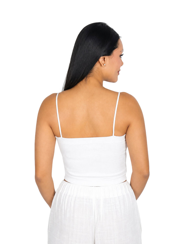 Mujer vistiendo blusa blanca corta de tirantes y pantalones blancos sobre fondo blanco.