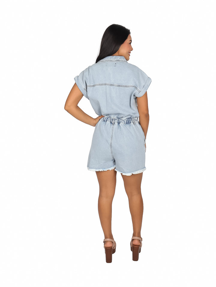 Mujer vistiendo un jumpsuit corto de mezclilla color azul claro con botones frontales y cintura elástica.