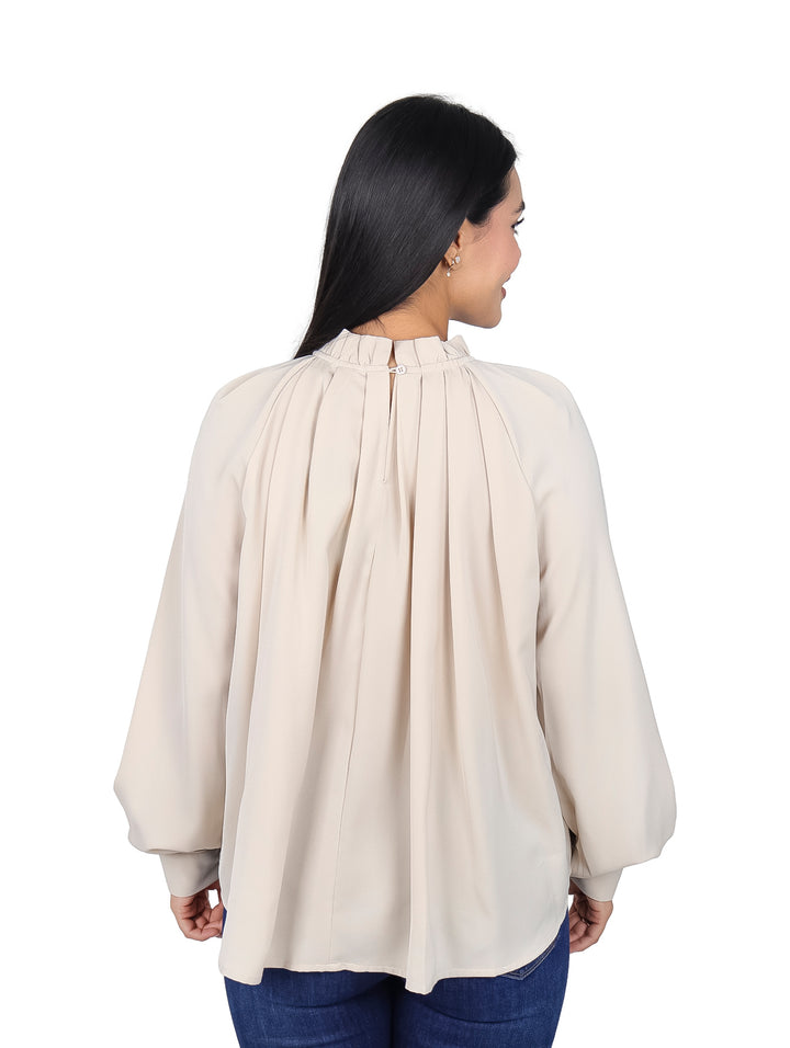 Mujer vistiendo blusa color beige de manga larga con cuello alto y pliegues frontales.