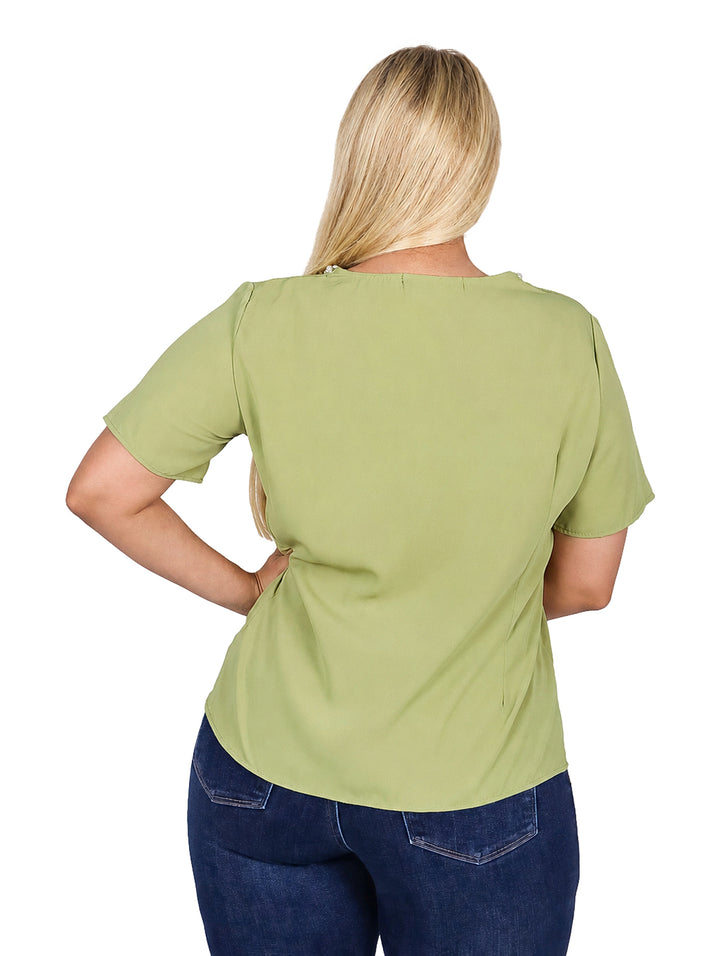 Mujer vistiendo blusa verde de manga corta con detalles de perlas en el cuello y jeans azules.