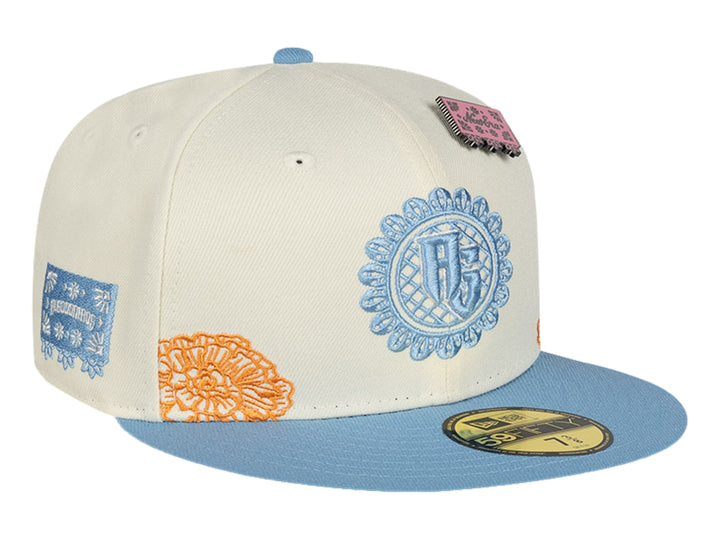Gorra New Era blanca con visera azul, detalles bordados en naranja y azul, y un pequeño parche rosa.