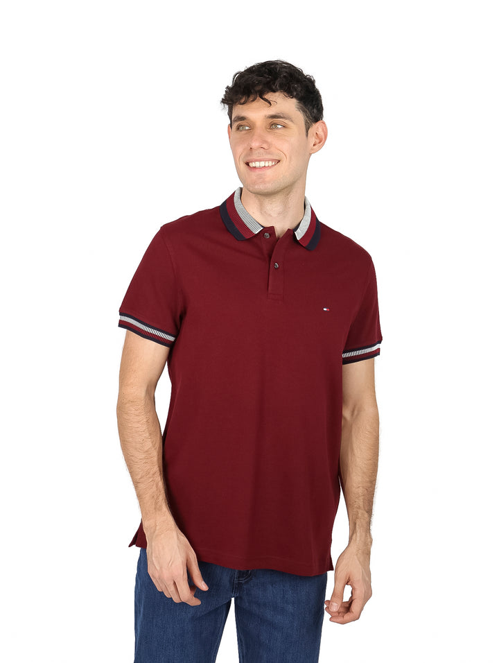 Hombre vistiendo una playera polo Tommy Hilfiger de color rojo oscuro con detalles en el cuello y mangas.