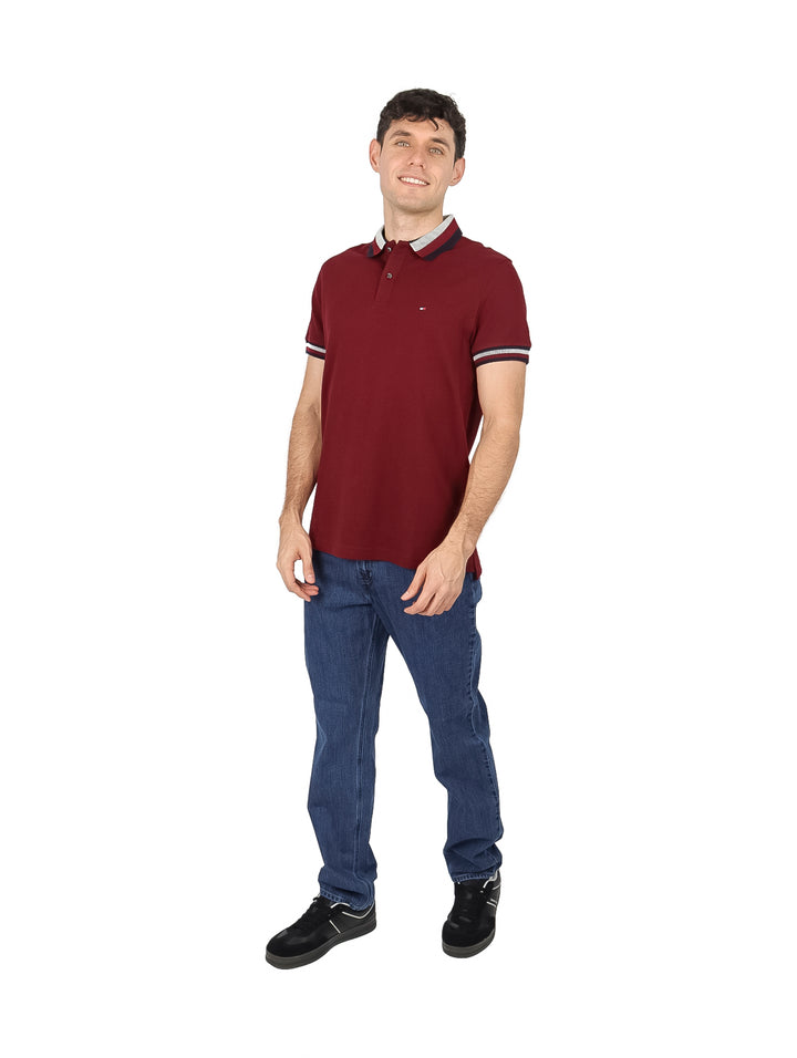 Hombre vistiendo una playera polo Tommy Hilfiger de color rojo oscuro con detalles en el cuello y mangas.