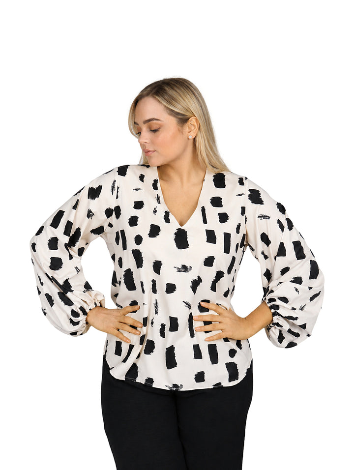 Mujer vistiendo blusa color beige con estampado abstracto negro, cuello en V y mangas largas acampanadas.