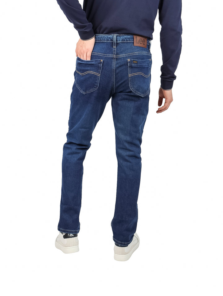 Hombre vistiendo pantalones LEE 112 slim fit en color azul stone con calzado blanco.