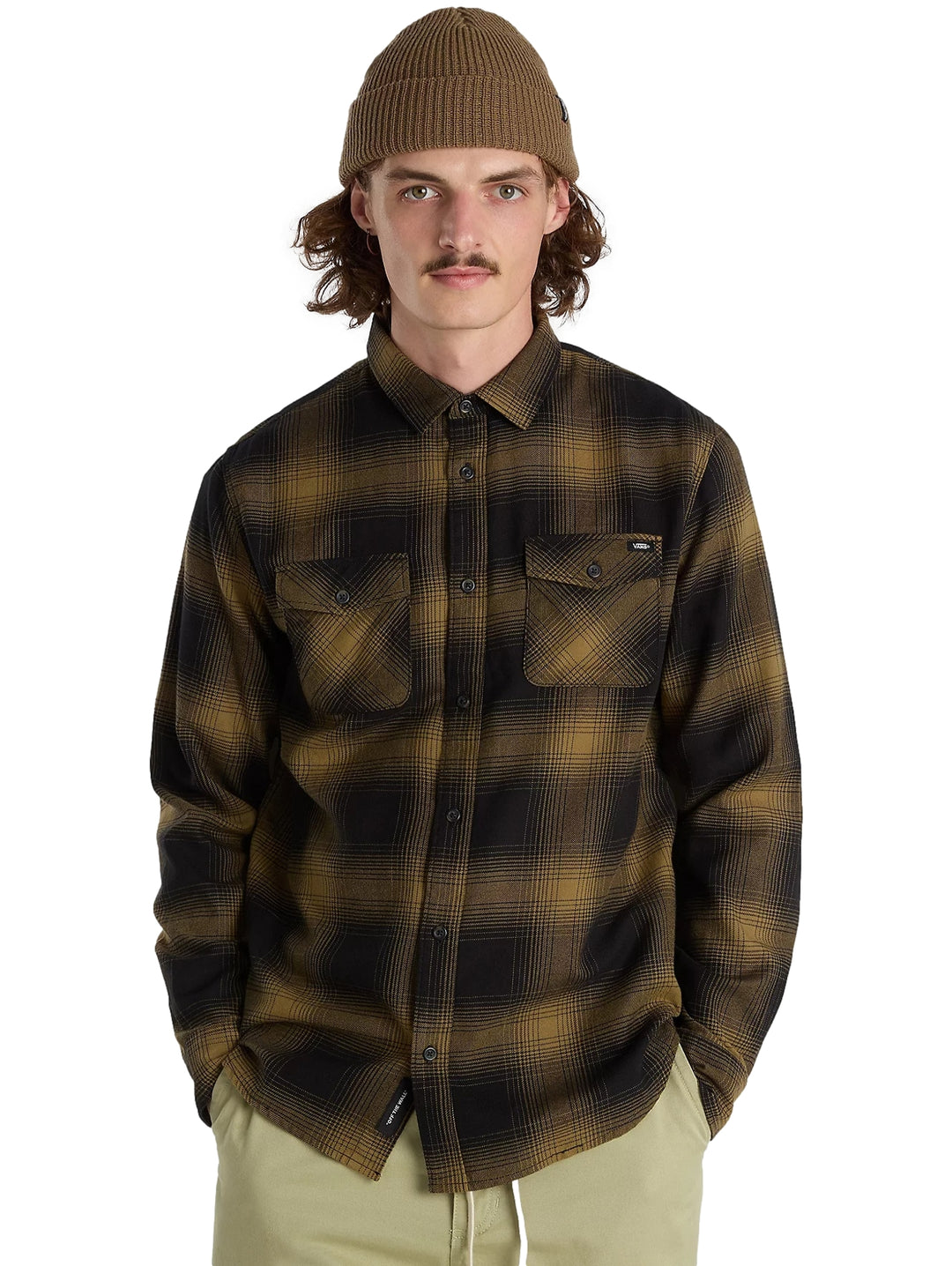 Hombre vistiendo camisa de franela a cuadros en colores negro y verde olivo, gorro marrón y pantalones beige.