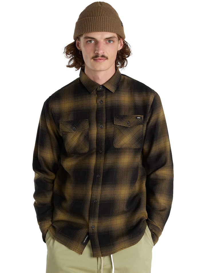 Hombre vistiendo camisa de franela a cuadros en colores negro y verde olivo, gorro marrón y pantalones beige.