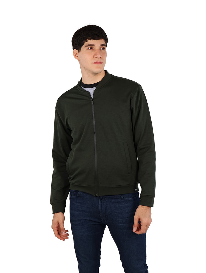 Hombre vistiendo una chamarra bomber color verde militar con cierre frontal y pantalones azul marino.
