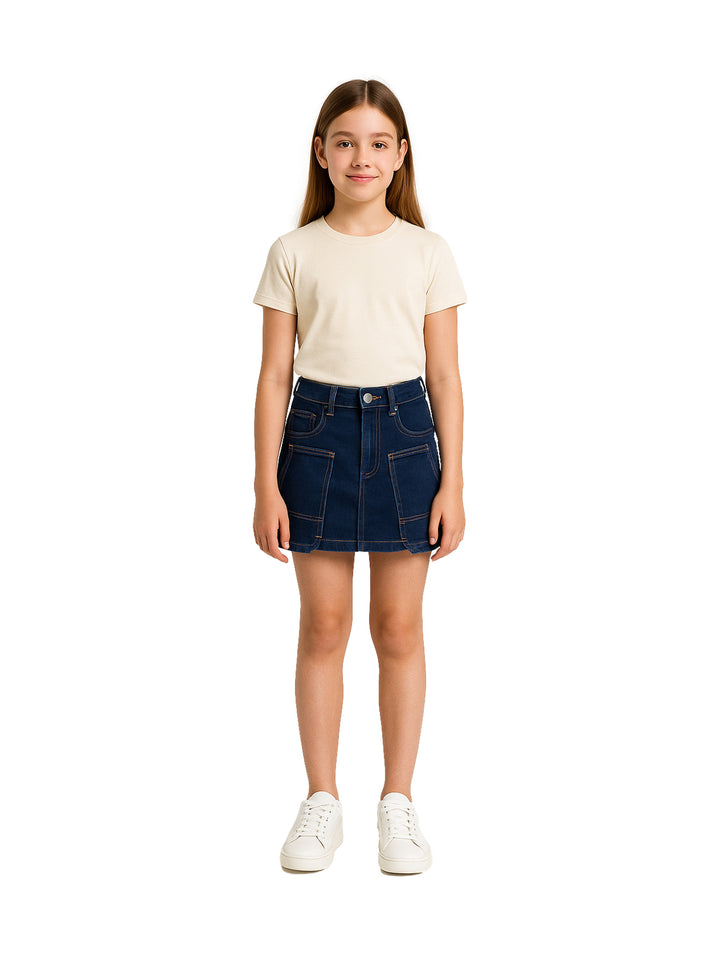 Niña vistiendo una falda corta de mezclilla azul oscuro estilo cargo con camiseta beige y tenis blancos.