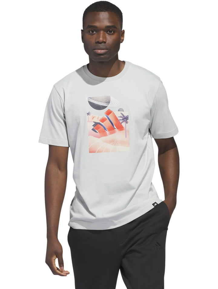 Hombre vistiendo camiseta blanca Adidas con estampado gráfico de paisaje playero en tonos naranja y azul.