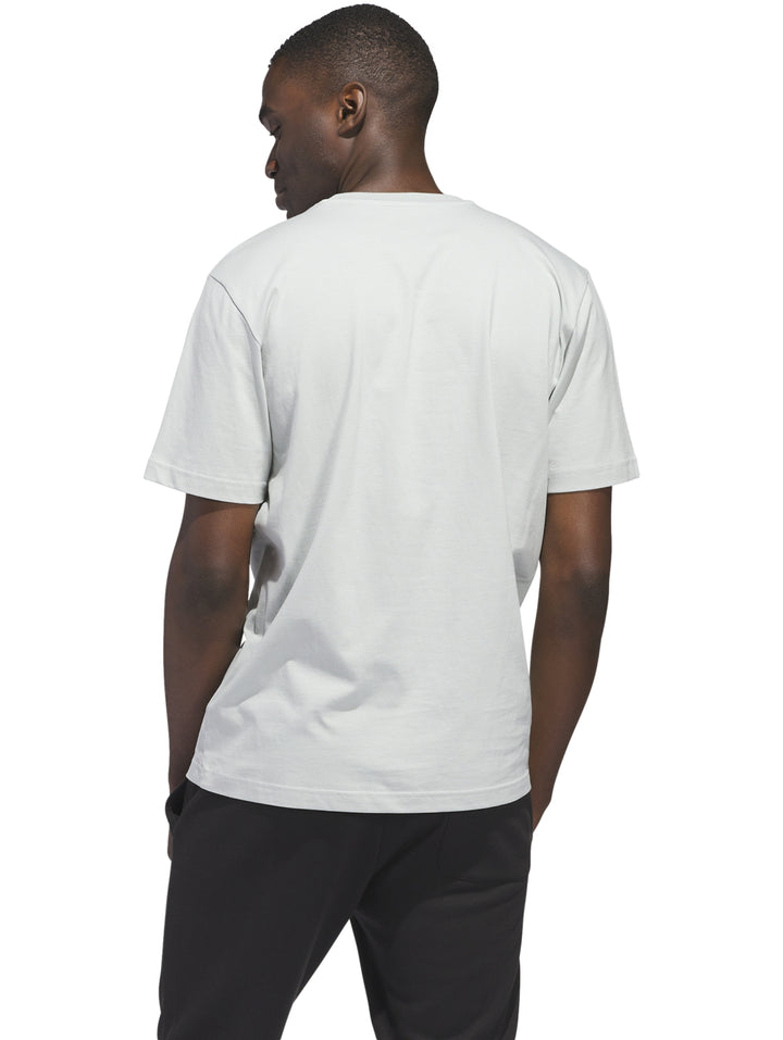 Hombre vistiendo camiseta blanca Adidas con estampado gráfico de paisaje playero en tonos naranja y azul.
