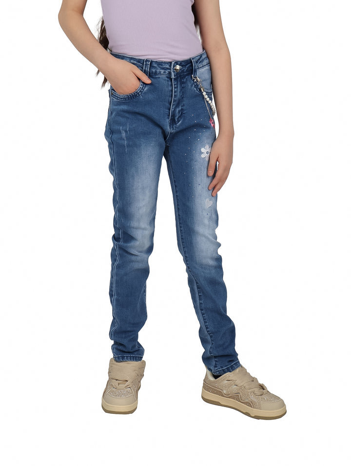 Pantalón de mezclilla azul para niña con detalles de flores blancas y una cadena decorativa lateral.