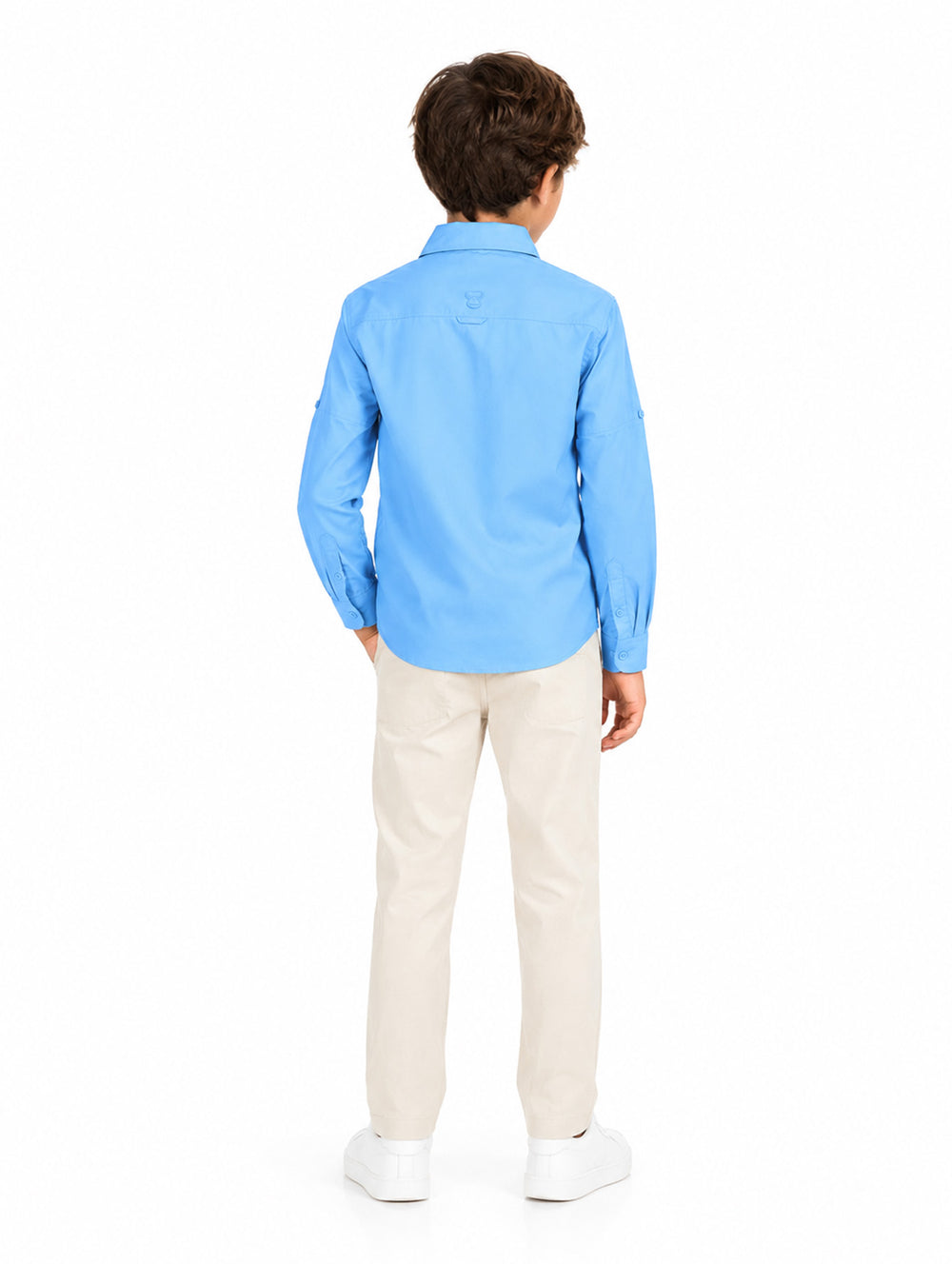 Ni�o modelando una camisa azul de manga larga, pantalones beige y tenis blancos sobre fondo blanco.