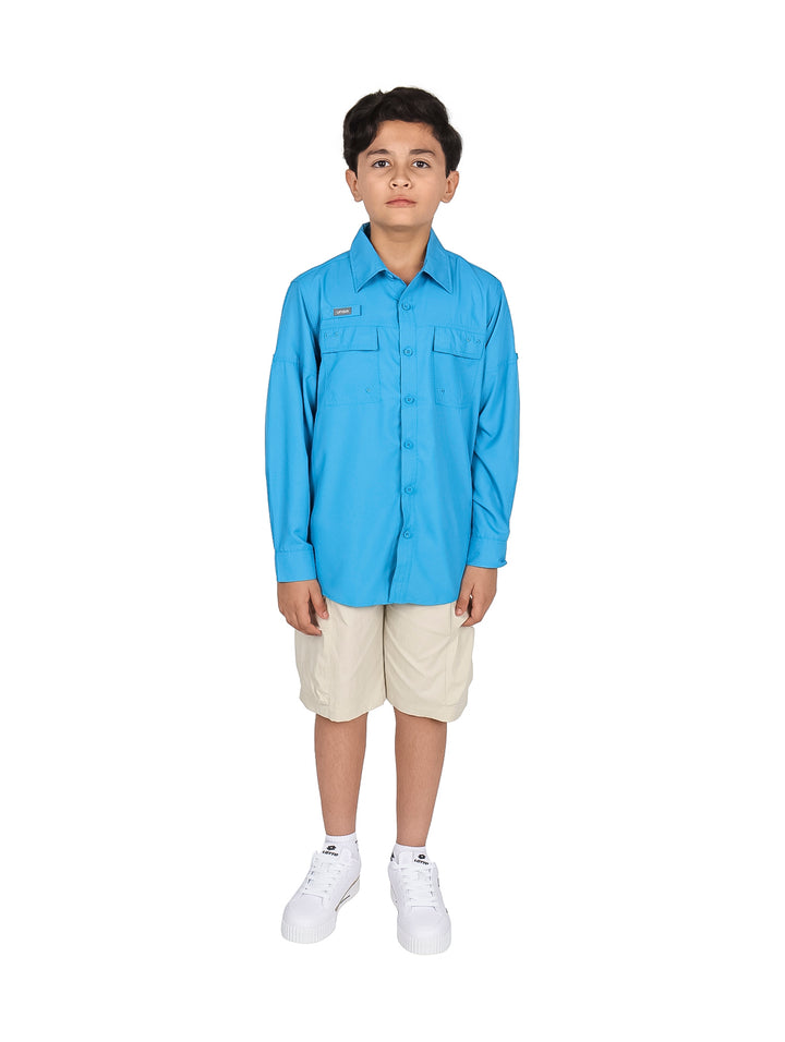 Niño modelando una camisa azul de manga larga, pantalones beige y tenis blancos sobre fondo blanco.