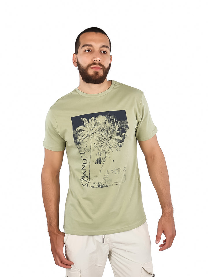 Hombre vistiendo una camiseta de manga corta color verde claro con un estampado frontal de palmeras en tonos azul y blanco.