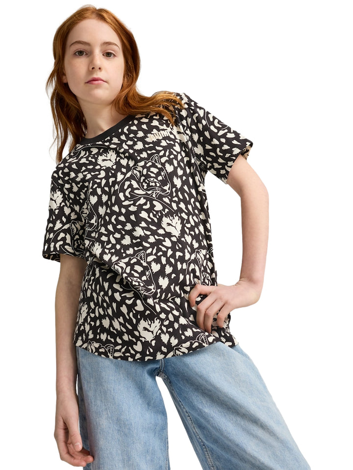 Niña vistiendo playera PUMA negra con estampado blanco all-over y pantalones vaqueros azules.
