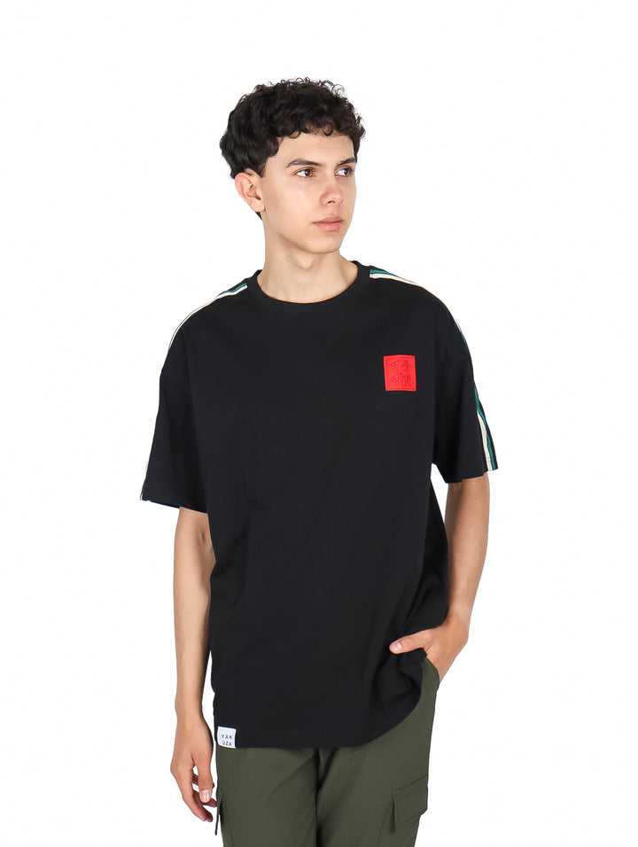 Hombre vistiendo playera YAKUZA color negro con un parche rojo en el pecho y detalles verdes en los hombros.