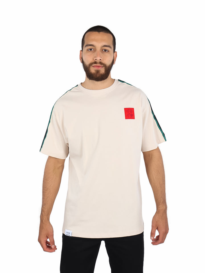Playera color beige de manga corta con rayas verdes en los hombros y un parche rojo en el pecho.