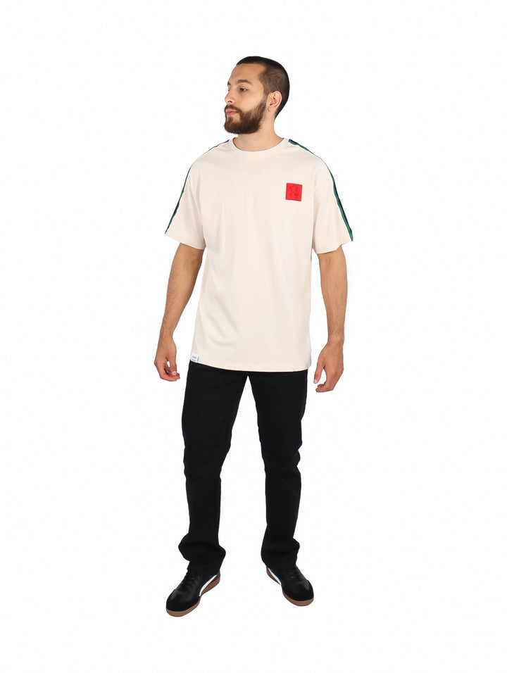 Playera color beige de manga corta con rayas verdes en los hombros y un parche rojo en el pecho.