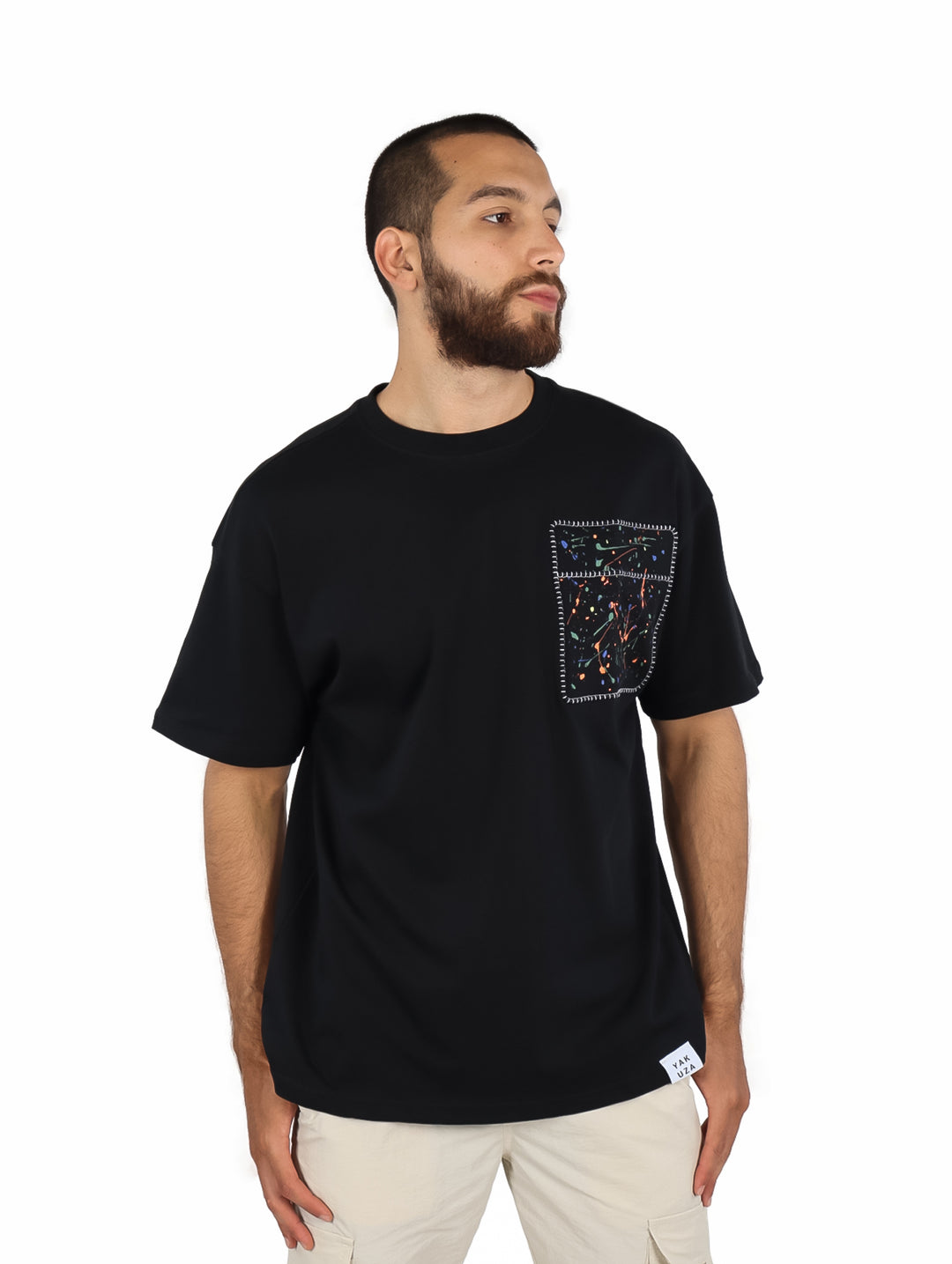 Hombre vistiendo playera negra de manga corta con un detalle decorativo cuadrado en el pecho izquierdo.
