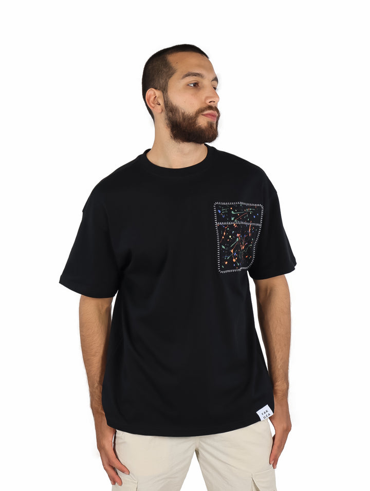 Hombre vistiendo playera negra de manga corta con un detalle decorativo cuadrado en el pecho izquierdo.
