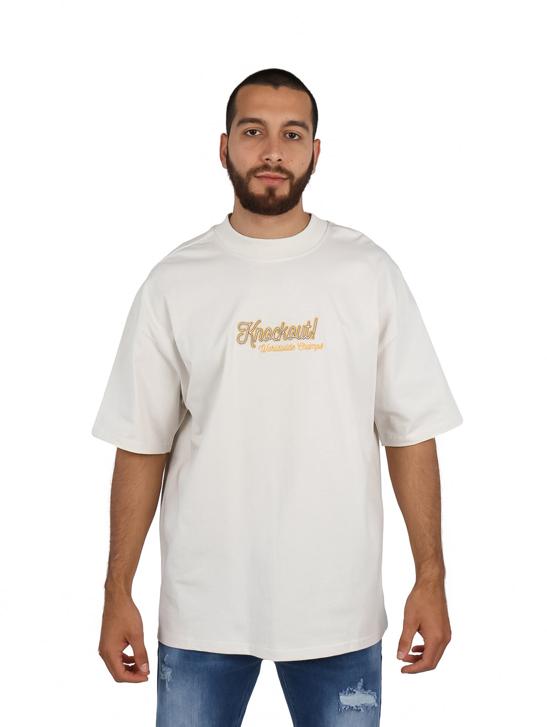 Hombre vistiendo playera oversize color arena con logo dorado en el pecho y pantalones mezclilla azules.